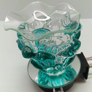 May include: A plug-in wax warmer featuring a clear, ruffled glass dish atop a textured, teal-colored glass base. The unit includes a circular mirrored platform, a black electrical plug housing, and a silver-toned control knob on the front.