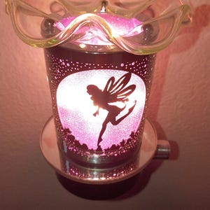 May include: A plug-in wax warmer featuring a cylindrical metallic body with a cutout silhouette of a fairy against a circular background. The interior emits a bright pink light. A clear, ruffled glass dish sits on top to hold wax melts.