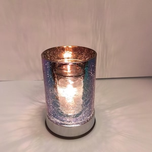 Puede incluir: Un portavelas cilíndrico de vidrio con textura agrietada y un acabado iridiscente multicolor que cambia entre tonos azules, morados y cobrizos. La base es de metal plateado reflectante y una vela encendida brilla en el interior.