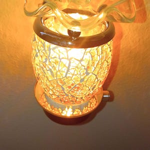 May include: A plug-in wax warmer featuring a glowing, amber-colored mosaic glass body with a cracked texture. The top consists of a clear, ruffled glass dish designed to hold wax, illuminated from within to cast a warm, golden light outward.