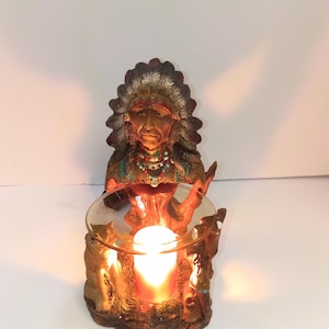 May include: A decorative resin figurine featuring a person wearing a traditional feathered headdress and beaded necklace, positioned behind a clear glass bowl. A glowing orange candle sits inside the bowl, illuminating the sculpted base with wolf figures.