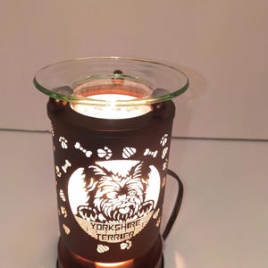 Puede incluir: Un calentador de cera eléctrico cilíndrico de color marrón oscuro con diseños recortados de huellas y huesos. El frente muestra un recorte circular de un Yorkshire Terrier con el texto "YORKSHIRE TERRIER" debajo. Un plato de vidrio transparente descansa encima.