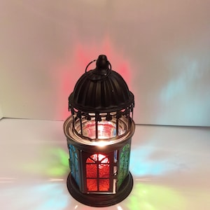 May include: A black metal lantern with a domed top and a circular hanging ring. The lantern features panels of textured glass in red, green, and blue, which cast colorful light reflections onto the surrounding area when illuminated from within.
