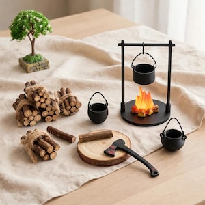 Peut inclure: Un ensemble de camping miniature comprenant un trépied en métal noir avec une marmite suspendue au-dessus d'un faux feu, deux marmites noires, des fagots de bois liés avec de la ficelle, une bûche, une petite hache et un arbre miniature.