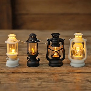 May include: Four miniature hurricane lanterns arranged in a row, featuring two white and two black models. Each lantern has a clear glass chimney, a metal handle, and an internal light source that emits a warm, glowing yellow illumination.