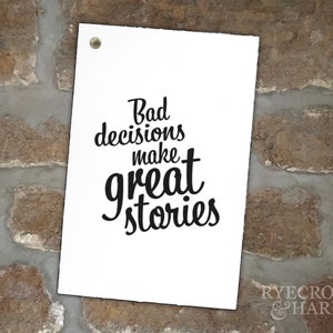 以下が含まれることがあります： 白い看板に黒い文字で「Bad decisions make great stories」と書かれています。看板はレンガの壁に掛けられています。
