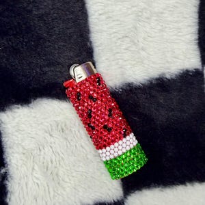 May include: A rectangular lighter fully covered in small, sparkling rhinestones arranged in a watermelon pattern. The design features a red body with scattered black rhinestones, a white horizontal band, and a green base with a metallic top.