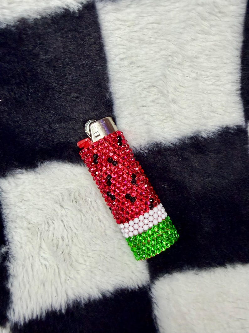 May include: A rectangular lighter fully covered in small, sparkling rhinestones arranged in a watermelon pattern. The design features a red body with scattered black rhinestones, a white horizontal band, and a green base with a metallic top.