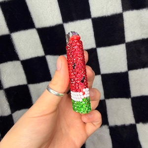 May include: A cylindrical lighter covered in small, sparkling rhinestones arranged in a watermelon pattern. The top section is red with scattered black stones, followed by a thin white band, and a green base. The metal ignition mechanism is visible.