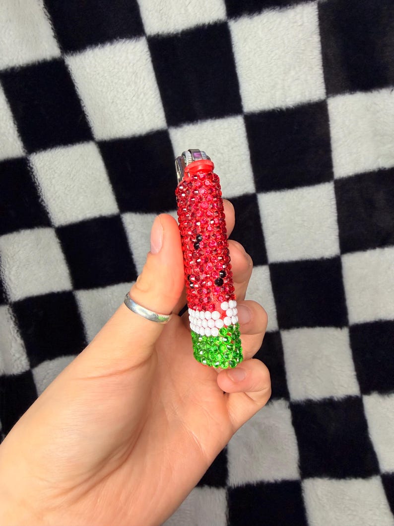 May include: A cylindrical lighter covered in small, sparkling rhinestones arranged in a watermelon pattern. The top section is red with scattered black stones, followed by a thin white band, and a green base. The metal ignition mechanism is visible.