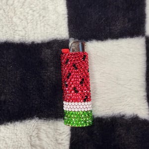 May include: A rectangular lighter covered in small, sparkling rhinestones arranged in a watermelon pattern. The top section features red stones with scattered black accents, followed by a thin white band, and a green base. A metallic ignition mechanism is visible.