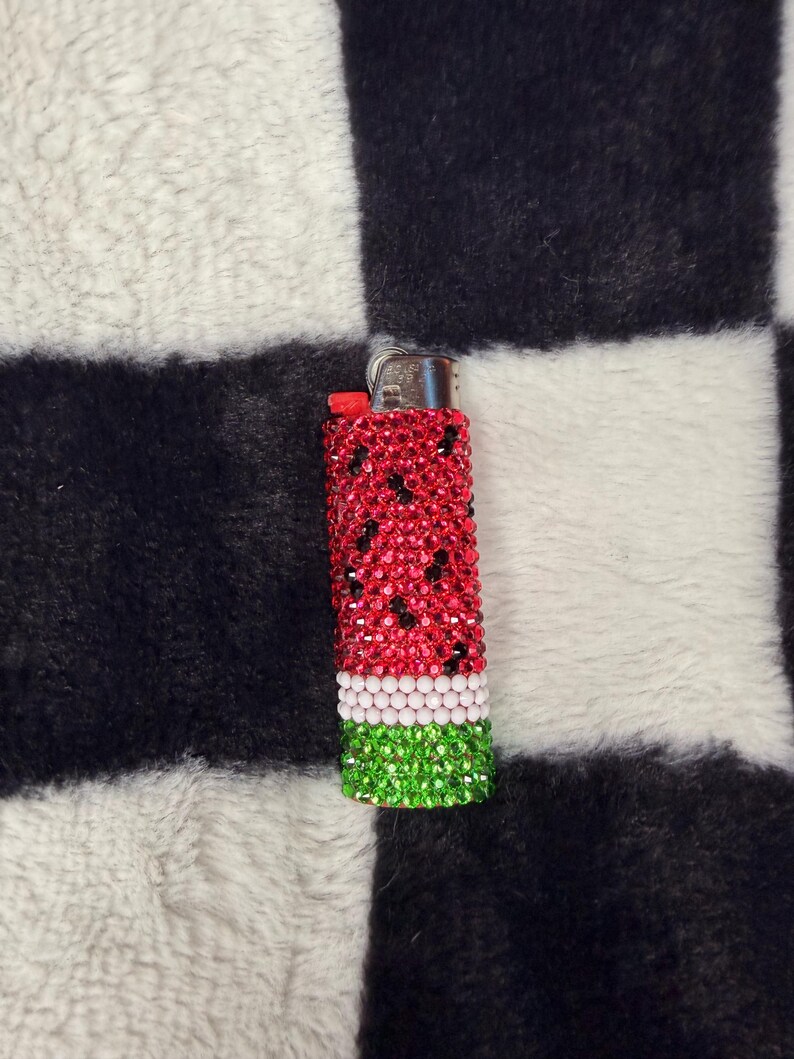 May include: A rectangular lighter covered in small, sparkling rhinestones arranged in a watermelon pattern. The top section features red stones with scattered black accents, followed by a thin white band, and a green base. A metallic ignition mechanism is visible.