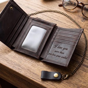 May include: A dark brown leather tri-fold wallet with a metal chain attachment. The interior features multiple card slots, a clear ID window, and an engraved message that reads, I love you more than you love your wallet, with brass snaps.