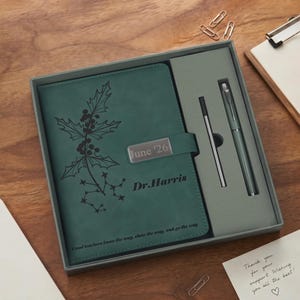 Pode incluir: Um caderno verde-escuro numa caixa cinzenta, com uma ilustração de um ramo de azevinho, o texto "June '26", "Dr. Harris" e "Good teachers know the way, show the way, and go the way". Inclui uma caneta a condizer e uma recarga de tinta fina.