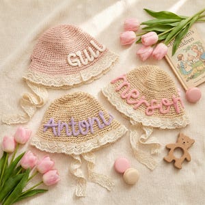 Puede incluir: Tres sombreros de paja con nombres personalizados en blanco, rosa y morado. Cada sombrero tiene un ribete de encaje y lazos de cinta. Los sombreros están dispuestos con tulipanes rosas, un libro infantil y macarons.