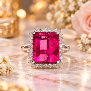Puede incluir: Un anillo con una gran piedra preciosa rectangular de color rosa vibrante montada en una banda de metal plateado. La piedra central está rodeada por un halo de pequeñas piedras blancas redondas y transparentes en un diseño festoneado.