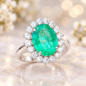 Puede incluir: Un anillo de tono plateado con una gran piedra preciosa de corte ovalado en color verde vibrante en el centro. La piedra central está rodeada por un halo de dieciséis pequeñas piedras blancas de corte redondo en una montura festoneada.