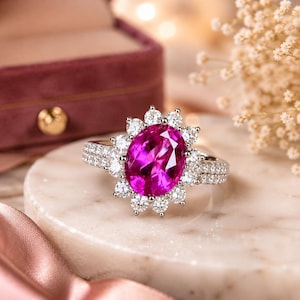 Op de afbeelding: Een zilverkleurige ring met een grote, ovaal geslepen, felroze middensteen omringd door een halo van kleine, ronde, helderwitte steentjes. De ringband is gespleten en aan de zijkanten ingelegd met extra kleine, helderwitte steentjes.