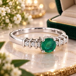 Op de afbeelding: Een zilverkleurige ring met een centrale rondgeslepen groene smaragd, geflankeerd door drie kleine rondgeslepen helderwitte diamanten aan elke kant, gezet in een gepolijste metalen band met een klauwzetting voor de centrale edelsteen.
