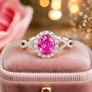 Puede incluir: Un anillo con una piedra preciosa rosa vibrante de corte ovalado en el centro, rodeada por un halo de pequeños diamantes blancos redondos. La banda está hecha de un material metálico y muestra un delicado patrón entrecruzado con diamantes.