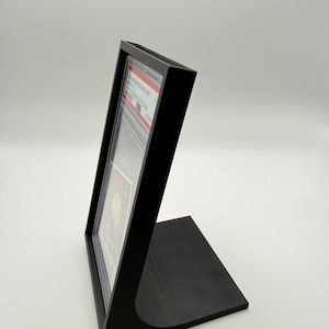 May include: A black display stand designed to hold a graded trading card. The stand is angled and has a rectangular base. The card is visible inside a clear protective case.