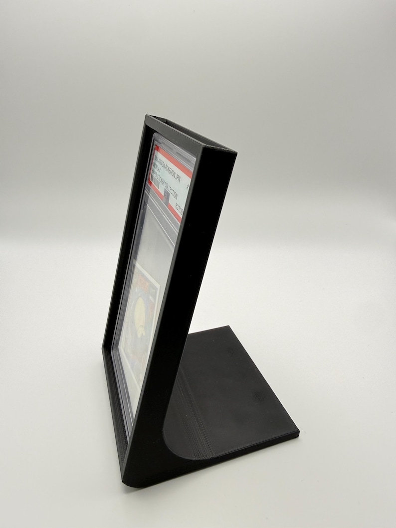 May include: A black display stand designed to hold a graded trading card. The stand is angled and has a rectangular base. The card is visible inside a clear protective case.