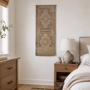 May include: A rectangular wall hanging featuring a beige background with dark brown geometric patterns and subtle red accents. The textile displays a symmetrical design with diamond shapes and Greek key motifs, mounted vertically on a plain white wall.