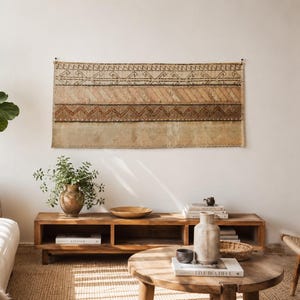 May include: A rectangular wall hanging featuring horizontal bands of geometric patterns in shades of brown, tan, and cream. The textile displays intricate zigzag and linear motifs across its surface, suspended horizontally against a plain wall.