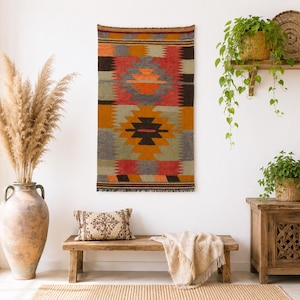 May include: A rectangular woven wall hanging featuring a geometric pattern in shades of red, orange, gray, and black. The textile displays symmetrical diamond motifs and horizontal bands, finished with a fringed edge at the top and bottom.