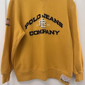 May include: Mustard yellow sweatshirt with the text "POLO JEANS COMPANY" in navy blue and white. The sweatshirt has a crew neck and long sleeves. The American flag is on the left sleeve.