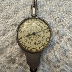Puede incluir: Un curvímetro de metal plateado con una esfera circular que presenta múltiples escalas concéntricas etiquetadas como statute miles, kilometres y nautical miles. El dispositivo tiene una pequeña rueda en la base y una anilla metálica.
