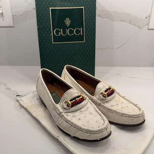 Peut inclure: Une paire de mocassins en cuir blanc texturé avec un mors en métal doré sur une bande rayée rouge et bleue. Les chaussures sont présentées avec un sac de protection en tissu blanc et une boîte verte haute marquée Gucci avec un logo doré.