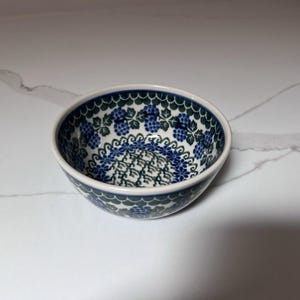 Puede incluir: Un cuenco de cerámica con un estampado floral azul y verde. El cuenco tiene un interior blanco y un borde festoneado. El diseño presenta racimos de uvas azules y hojas verdes.
