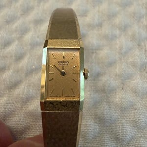 May include: A gold-tone Seiko Quartz watch with a rectangular face and a textured gold dial. The watch features thin gold hour markers, a matching gold-tone metal band with a brushed finish, and a small crown on the right side of the case.