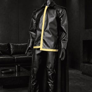 Peut inclure: Un mannequin noir portant une veste noire à manches longues avec une bande verticale jaune le long de la fermeture avant et un ourlet jaune, assortie d'un pantalon noir et d'une longue cape noire tombant des épaules jusqu'au sol.