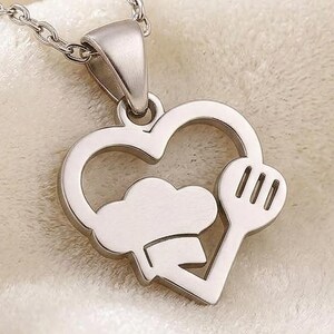 May include: A silver-toned metal necklace featuring a heart-shaped pendant. The interior of the heart cutout contains a stylized chef hat and a slotted spatula. The pendant is attached to a delicate silver-toned chain with small circular links.