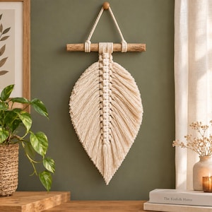 May include: A cream-colored macrame wall hanging shaped like a large leaf, suspended from a horizontal wooden branch by a thick cord. The fiber art features intricate knotted patterns along the central spine with long, frayed fringe edges.