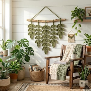 May include: A wall hanging featuring five crocheted olive green leaf strands suspended from a natural wooden branch. The leaves are arranged in a symmetrical pattern, creating a botanical display that hangs against a white horizontal paneled wall.