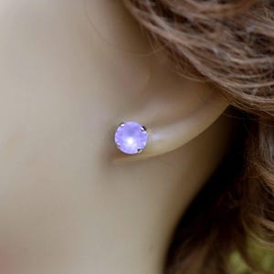 May include: A close-up of a light purple stud earring. The earring is round with a silver setting and is set against the backdrop of a person's ear and hair.