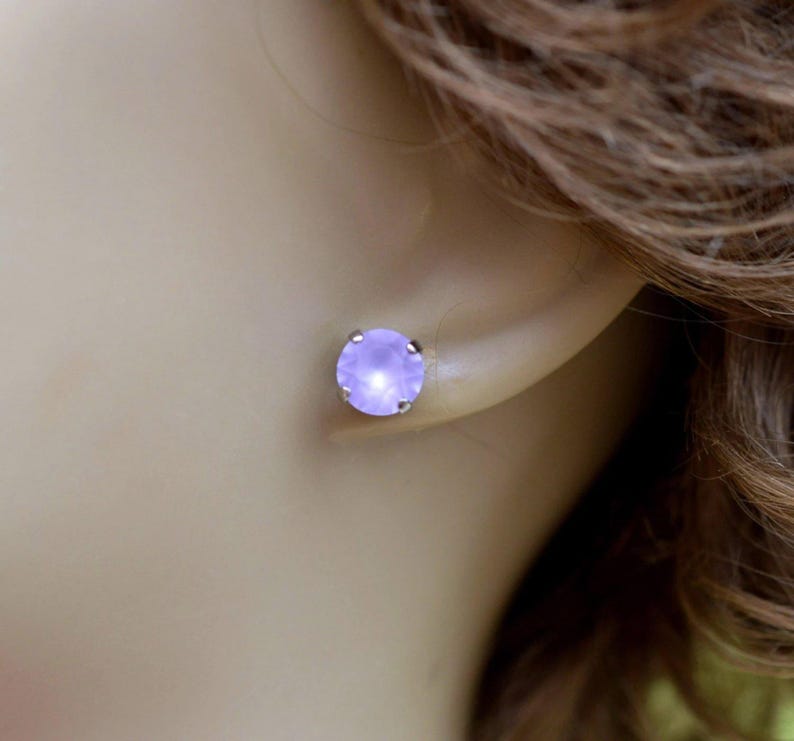 May include: A close-up of a light purple stud earring. The earring is round with a silver setting and is set against the backdrop of a person's ear and hair.