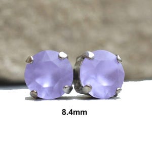 May include: A pair of stud earrings featuring round, faceted lavender-colored stones. Each stone is held in place by four silver-toned prongs. The text "8.4mm" is displayed below the earrings, indicating the size of the stones.