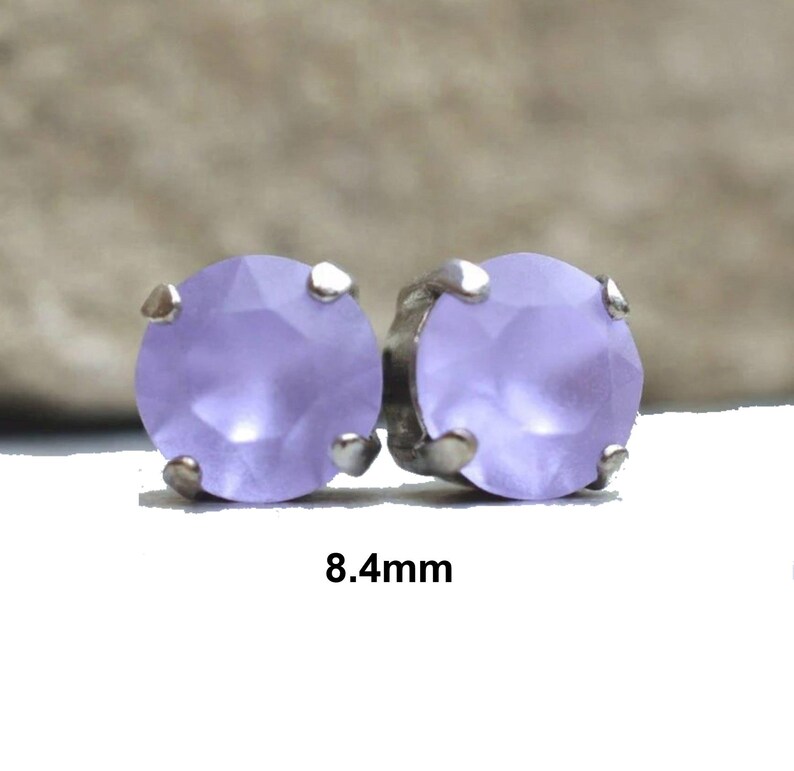 May include: A pair of stud earrings featuring round, faceted lavender-colored stones. Each stone is held in place by four silver-toned prongs. The text "8.4mm" is displayed below the earrings, indicating the size of the stones.