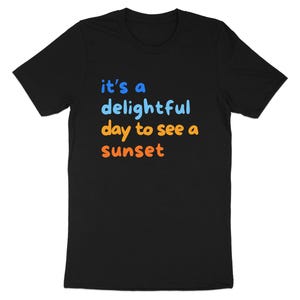 May include: A black short-sleeved t-shirt featuring the text "it's a delightful day to see a sunset" printed on the front. The words "it's a" and "delightful" are in light blue, while "day to see a" and "sunset" are in orange.