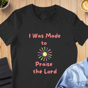 May include: A black short-sleeved t-shirt featuring the pink text "I Was Made to Praise the Lord" centred on the chest. Below the word "to" is a graphic of a yellow sun with multi-coloured rays in shades of pink, purple, blue, green, and orange.
