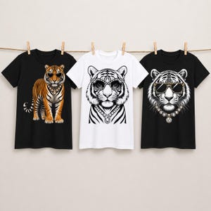 Könnte beinhalten: Drei kurzärmelige T-Shirts hängen an einer Leine. Links ein schwarzes T-Shirt mit einem orangefarbenen Tiger mit Sonnenbrille und Halskette. In der Mitte ein weißes T-Shirt mit einem Tigergesicht mit Leopardenbrille und Kette. Rechts ein schwarzes T-Shirt mit einem Tigergesicht mit Leopardenbrille und Kette.
