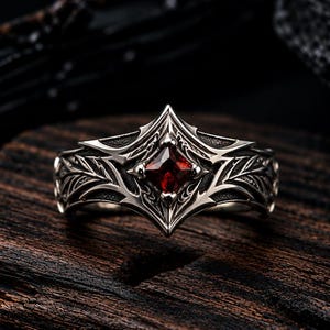 May include: A silver ring featuring a central square-cut deep red gemstone set within a pointed, ornate metal frame. The band is decorated with intricate, symmetrical leaf-like engravings that extend from the center setting along the sides.