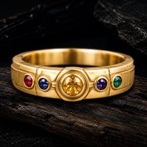 May include: A gold band ring featuring a central round yellow gemstone flanked by four smaller round stones in red, blue, purple, and green. The metal surface has a segmented, grid-like texture surrounding each of the five bezel-set circular gems.