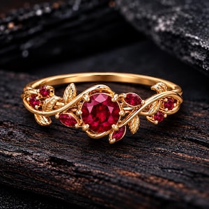 Könnte beinhalten: Ein goldener Ring mit einem zentralen, rund geschliffenen roten Edelstein, umgeben von kleineren roten Steinen und geformten Goldblättern. Die Ringschiene ist in einem Rankenmuster gestaltet und mit weiteren roten Akzenten verziert.