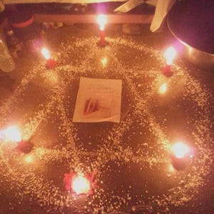 May include: A pentagram shape formed by white granular material on a dark surface, illuminated by five red candles placed at each point. A rectangular white paper with printed text and a small image is positioned in the center of the pentagram.