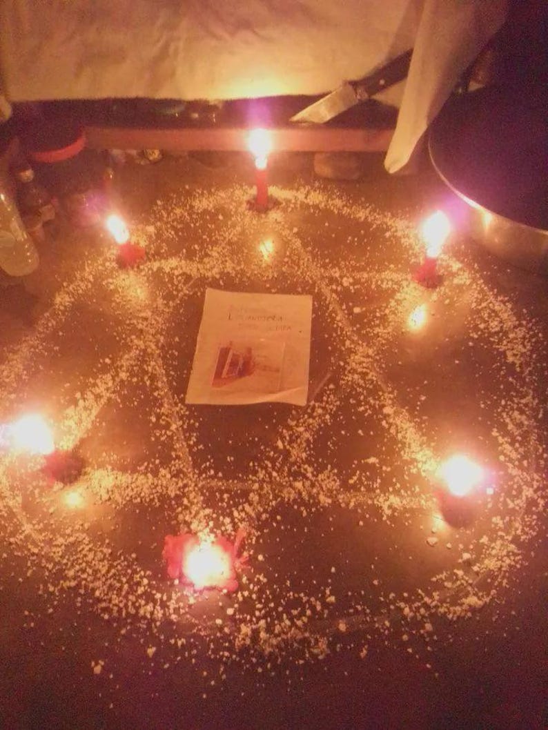 May include: A pentagram shape formed by white granular material on a dark surface, illuminated by five red candles placed at each point. A rectangular white paper with printed text and a small image is positioned in the center of the pentagram.